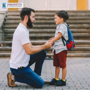A man kneels to talk to a smiling young boy with a backpack in front of outdoor steps. The Simonetti & Associates logo is visible in the top left corner.
