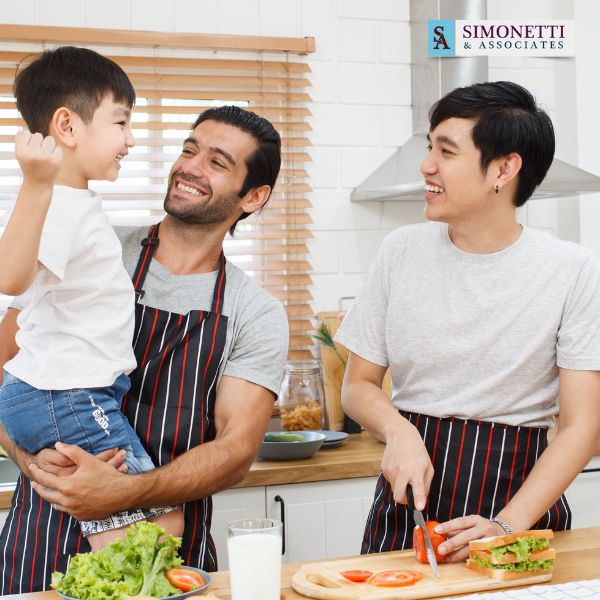 Two men in aprons prepare vegetables in a kitchen while smiling at a young boy, with a "Simonetti & Associates" logo in the top right corner.