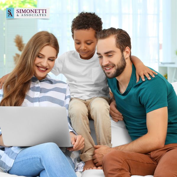 A woman, man, and young boy sit together on a couch, looking at a laptop. The Simonetti & Associates logo appears in the top left corner.