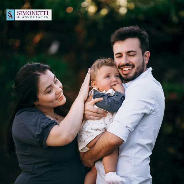 A smiling man holds a baby while a woman touches the baby’s head. The Simonetti & Associates logo is in the upper left corner.