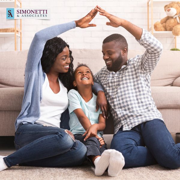 A smiling family sits on the floor, with two adults forming a roof shape over a child, in a living room. The Simonetti & Associates logo appears in the top left corner.