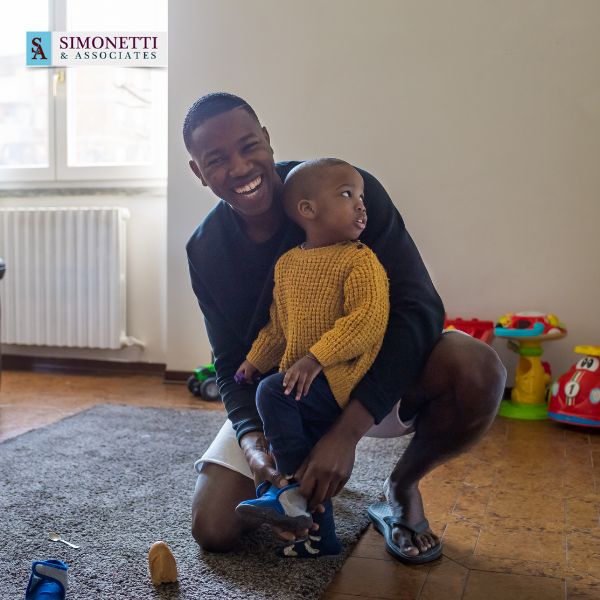 An adult helps a young child put on shoes in a playroom filled with toys and natural light from a window. The Simonetti & Associates child custody attorney logo appears in the top left corner.