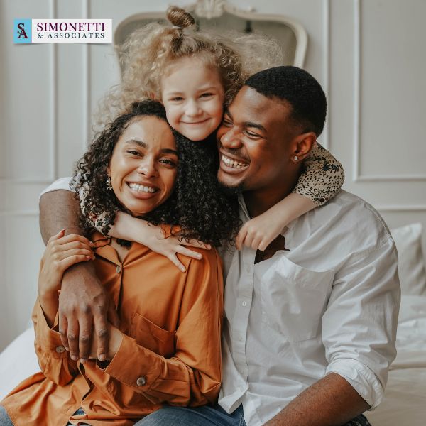 A smiling family of three sits close together on a bed, with a young girl hugging an adult woman and man. The Simonetti & Associates logo appears in the top left corner, showcasing your trusted Long Island Adoption Attorney.