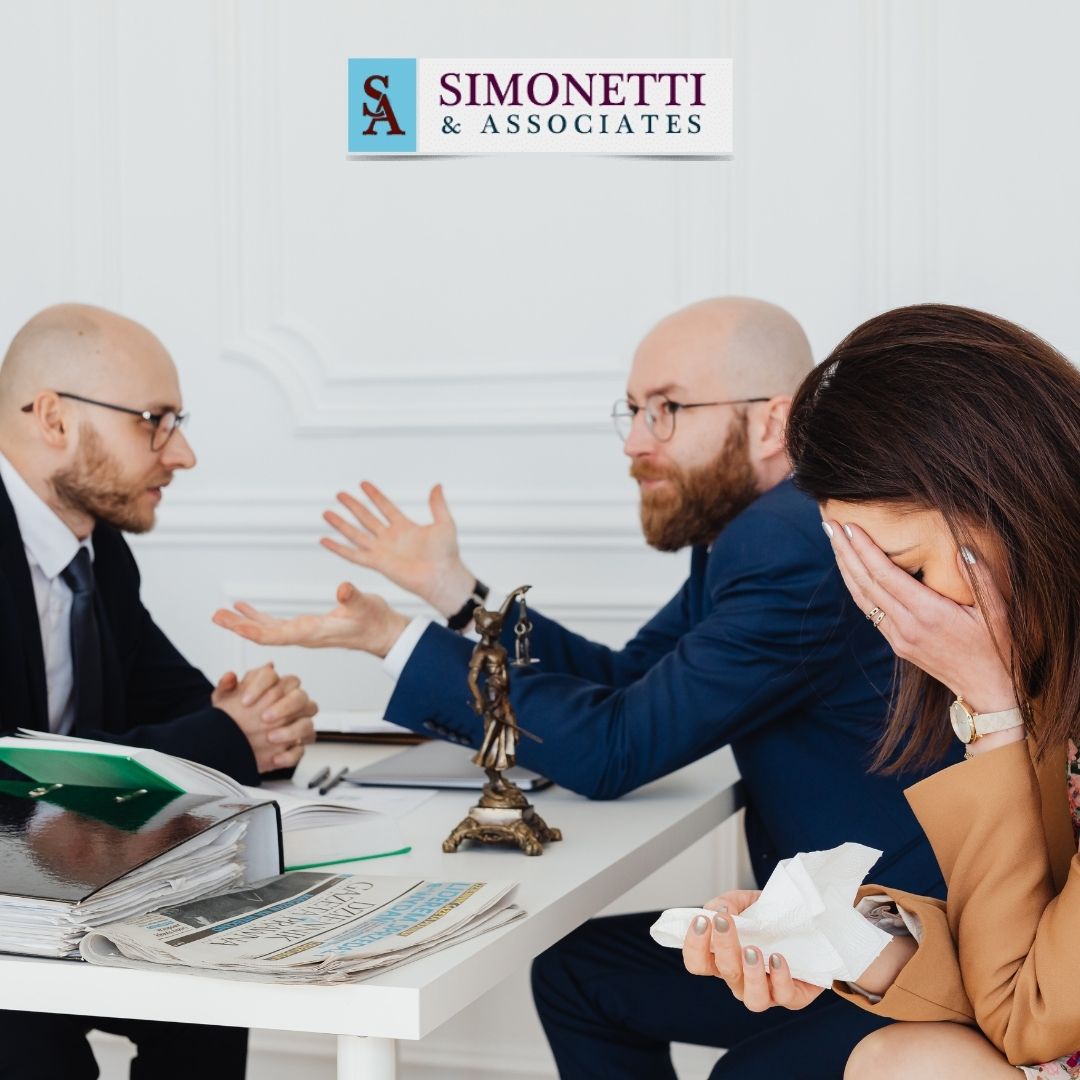 Three people sit at a table with documents; two men are talking while a woman, visibly upset, holds a tissue. A "Simonetti & Associates" sign on the wall suggests they may be meeting with a divorce lawyer in Massapequa.