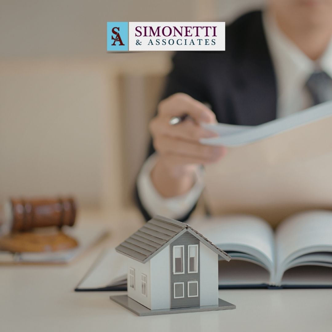 A person in a suit holding papers sits at a desk with a gavel, open book, and model house, symbolizing legal expertise; Simonetti & Associates logo at the top highlights their role as a trusted divorce lawyer on Long Island.
