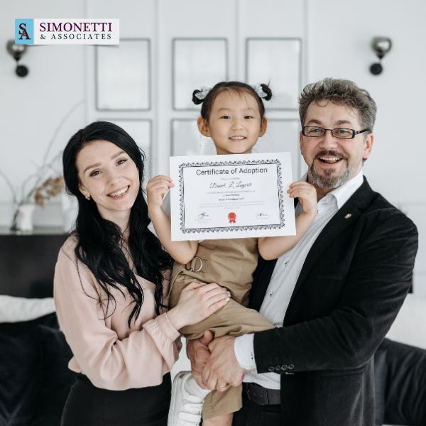 A smiling couple holds a young girl who displays a certificate of adoption, celebrating their new beginning. The logo "Simonetti & Associates" identifies them as trusted Nassau County divorce lawyers in the top left corner.