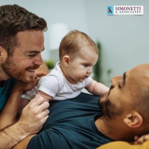 Two men smile and interact with a baby lying on one man’s chest. The Simonetti & Associates logo in the corner highlights their reputation as the best adoption lawyer on Long Island.