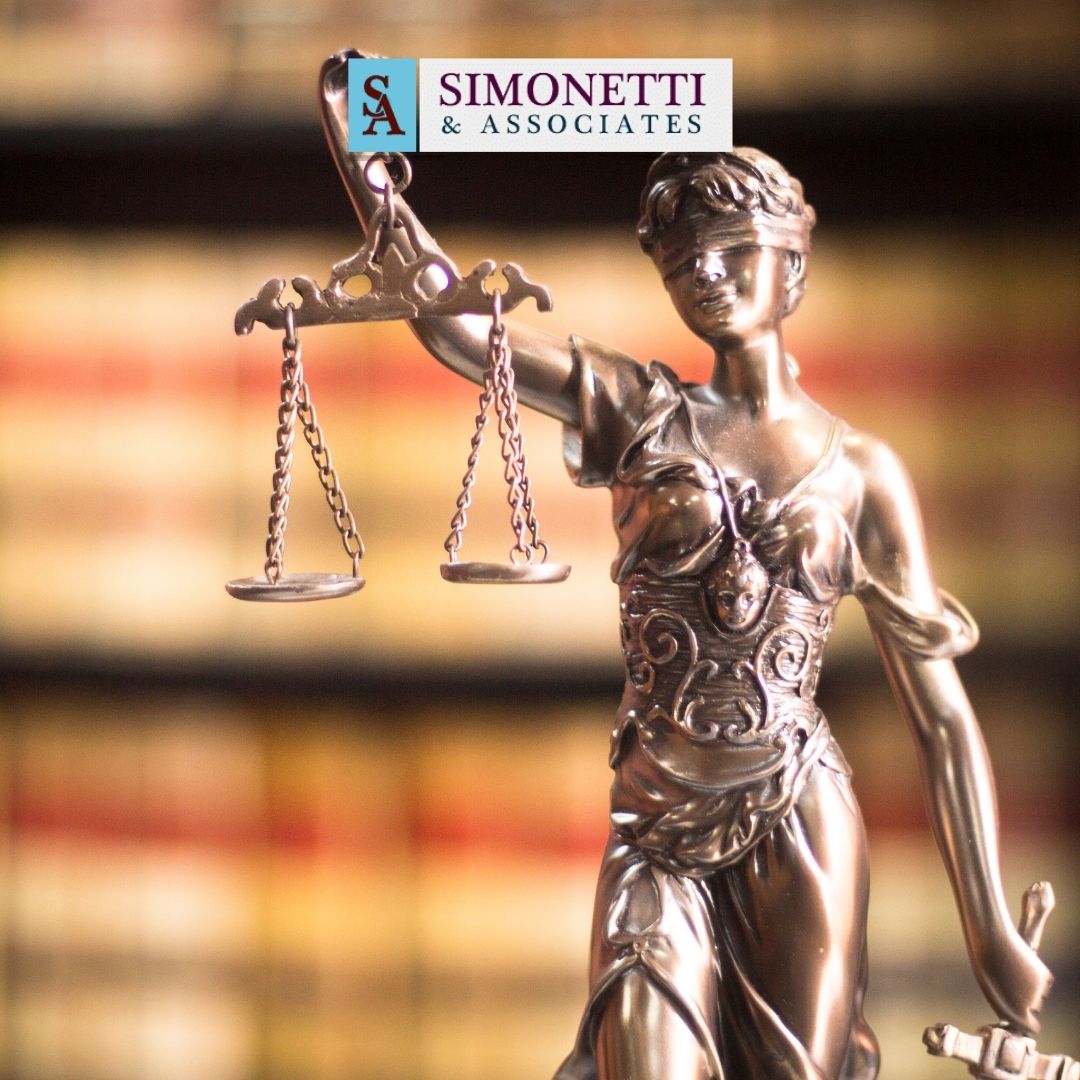 Bronze statue of Lady Justice holding scales, with the Simonetti & Associates logo above her and blurred bookshelves in the background, representing the best divorce lawyer on Long Island.