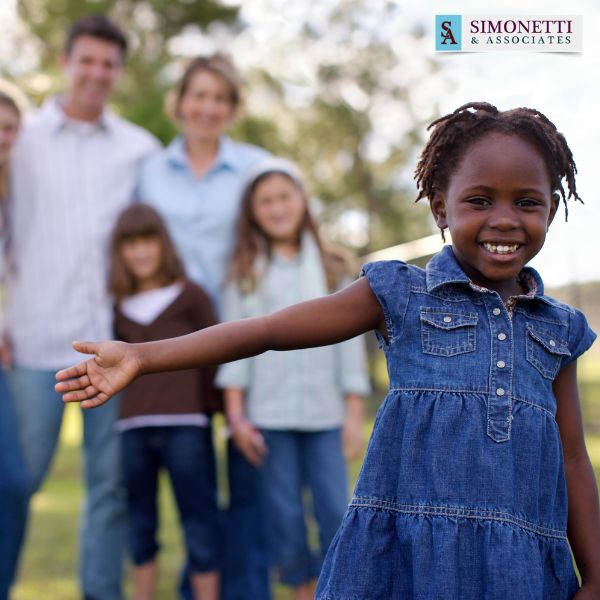 A young girl smiles and extends her arm toward the camera, with a group of adults and children in the blurred background. The Simonetti & Associates logo is visible in the corner, highlighting their role as a trusted Massapequa family divorce lawyer.