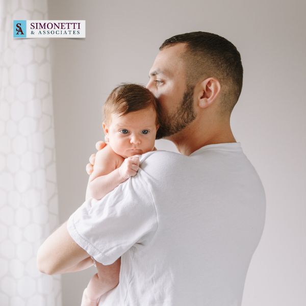 A man holds and kisses a baby on the head while standing by a window. The Simonetti & Associates logo, Divorce Lawyer in Hicksville, is visible in the top left corner.