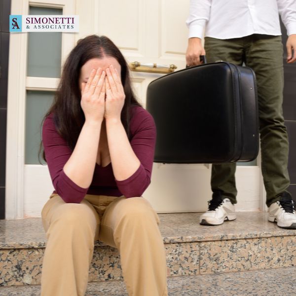 A woman sits on steps covering her face while a person stands nearby holding a suitcase. A Simonetti & Associates sign, known as the best divorce lawyer on Long Island, is visible on the wall.