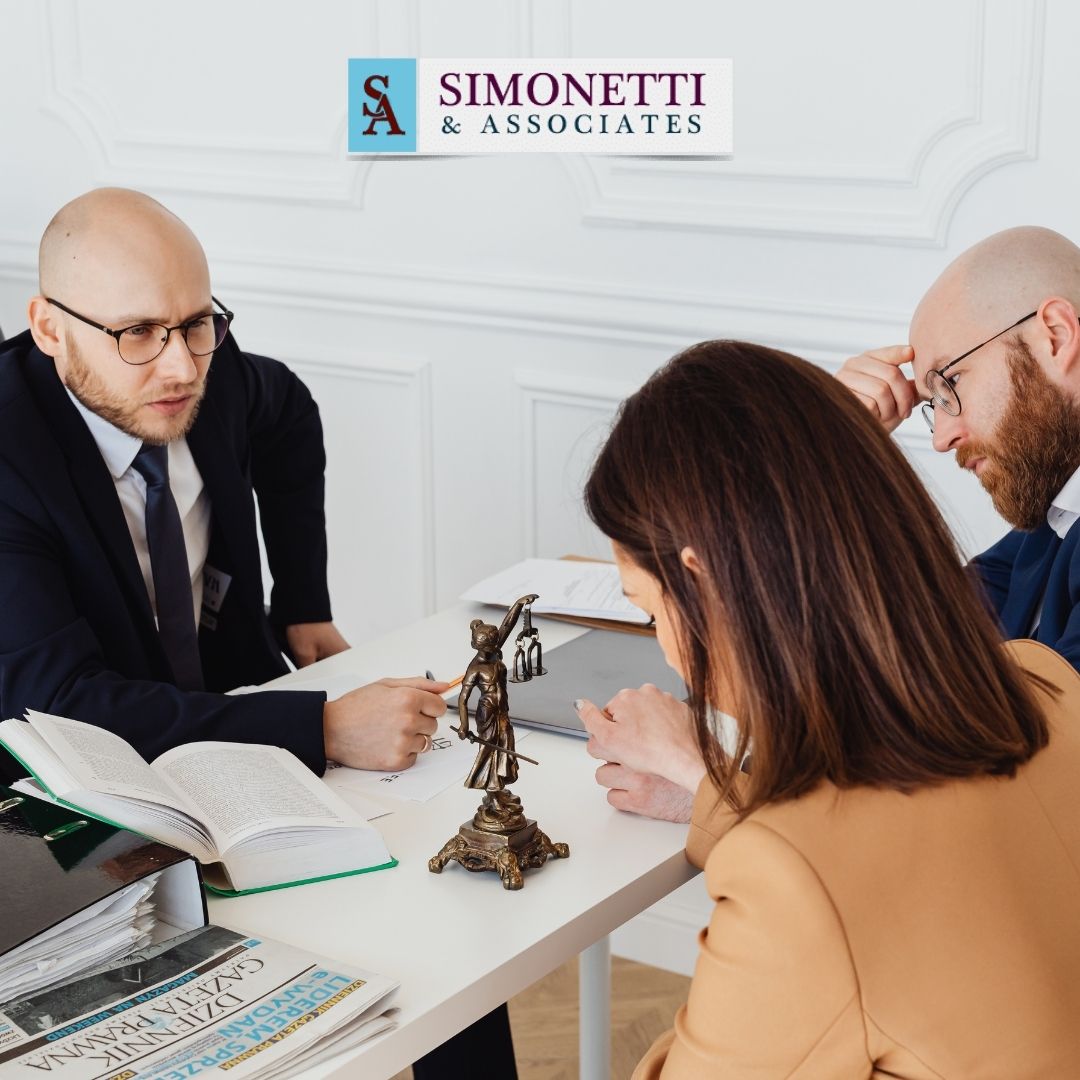 Three people in business attire sit around a table with legal books and a justice statue, discussing documents. The Simonetti & Associates logo is visible on the wall, highlighting their expertise as a top divorce attorney in Huntington.