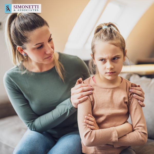 A woman sits beside a young girl with crossed arms who appears upset; the woman, perhaps seeking advice from a Huntington divorce lawyer, gently places her hands on the girl's shoulders, offering comfort.