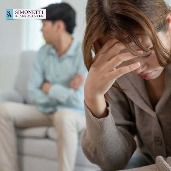 A woman sits with her head in her hand looking distressed, while a man sits in the background looking away; the Simonetti & Associates logo and Huntington divorce lawyer information are visible.