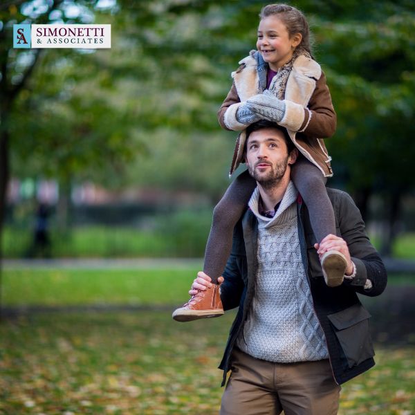 A man carries a smiling young girl on his shoulders in a park. The Simonetti & Associates logo appears in the top left corner, highlighting their trusted reputation as a leading divorce lawyer in Huntington.