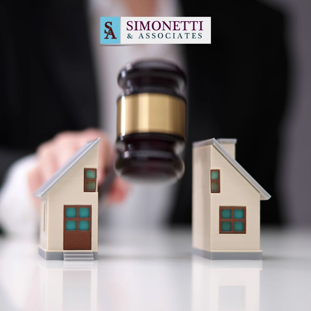A judge’s gavel comes down between two model houses, with a blurred figure in the background and a Simonetti & Associates logo above, highlighting the role of a Huntington divorce lawyer in property disputes.