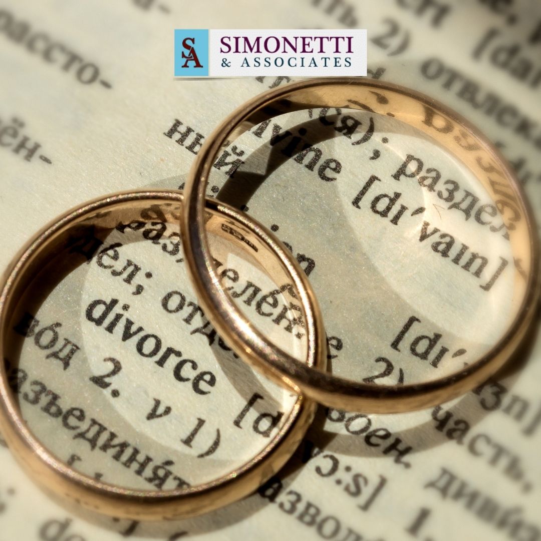 Two gold wedding rings rest on a dictionary page with the word "divorce" visible; at the top, the Simonetti & Associates logo highlights their role as a trusted divorce lawyer in Massapequa.