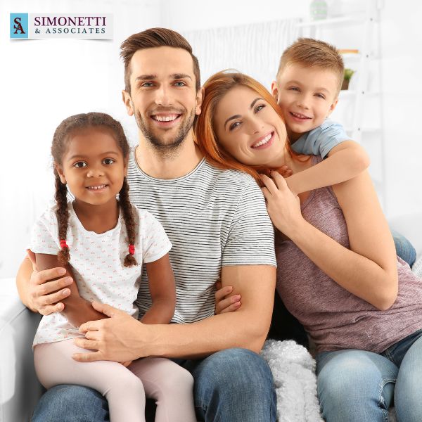 A smiling family of four, two adults and two children, sit closely together on a couch. The Simonetti & Associates logo in the top left corner highlights trusted divorce lawyer in South Hempstead services.