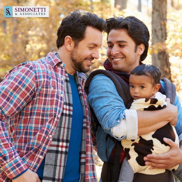 Two men smile at each other while one holds a baby wrapped in a blanket outdoors. The logo for Simonetti & Associates, trusted divorce lawyer in Manhasset, is visible in the top left corner.