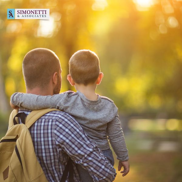 An adult carrying a child outdoors with sunlight in the background. The adult has a backpack and the child has an arm around their neck. "Simonetti & Associates," divorce lawyer in Glen Cove, logo is in the top left corner.