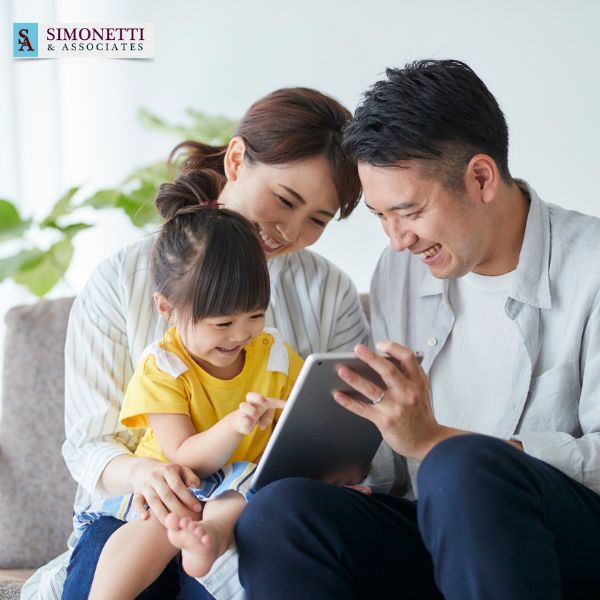 A smiling family of three sits on a couch, looking at a tablet together. The Simonetti & Associates logo is visible in the top left corner, showcasing the trusted Massapequa divorce lawyer's commitment to families.
