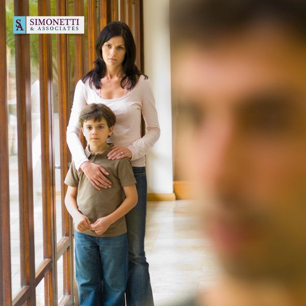 A woman stands behind a young boy, resting her hands on his shoulders, while a man appears blurred in the foreground. A Simonetti & Associates logo at the top left highlights their role as a trusted divorce lawyer in Massapequa.