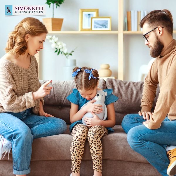 A woman and a man talk to a sad girl hugging a stuffed animal on a couch. The logo "Simonetti & Associates" is visible in the top left corner, suggesting support from a trusted divorce lawyer in Northport.