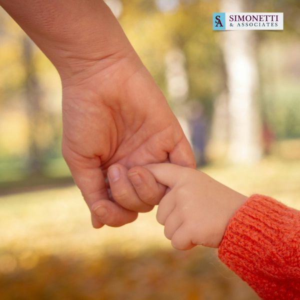 An adult holding a child's hand outdoors, with autumn colors in the background and a Simonetti & Associates logo in the corner, highlighting trusted Divorce Lawyer in Smithtown services.