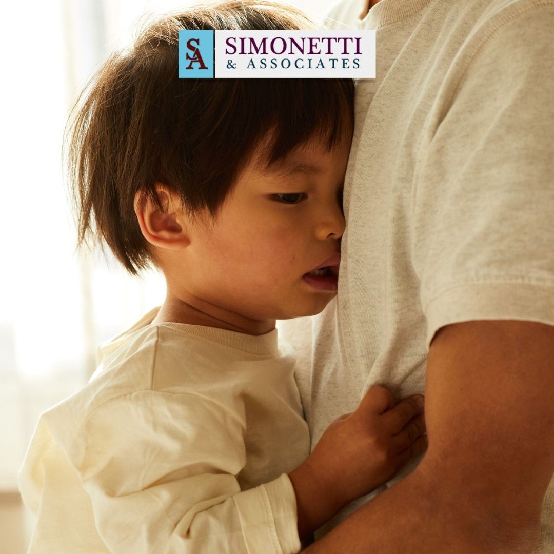 A young child hugs an adult's torso indoors, reflecting the emotional side of family matters. The logo "Simonetti & Associates," a trusted divorce attorney in Nassau County, appears at the top of the image.