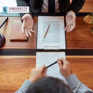 Two people sit across a wooden desk with documents; one gestures while the other holds a pen. A legal scale and the Simonetti & Associates logo highlight their discussion with an experienced Nassau Divorce Lawyer.