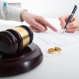 A person signs a document beside a judge’s gavel and two gold wedding rings, with the Simonetti & Associates logo in the corner, highlighting the trusted guidance of a Suffolk Divorce Lawyer.