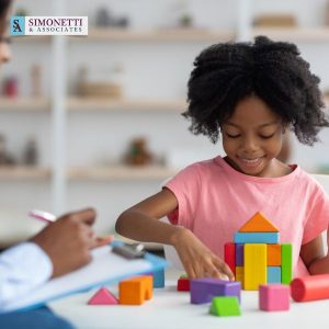 A young girl builds a colorful structure with wooden blocks while an adult, possibly a Divorce Lawyer On Long Island, takes notes in the background; Simonetti & Associates logo is visible.