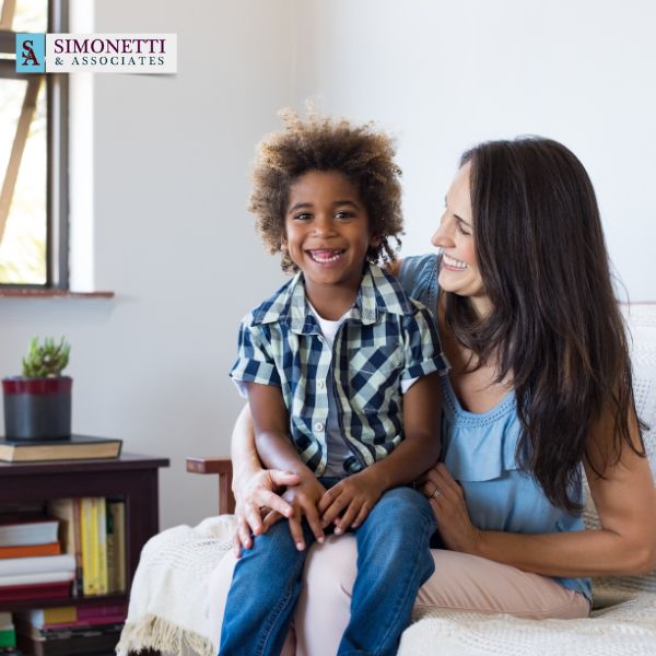 A woman and a child sit on a couch smiling together. The logo for Simonetti & Associates, Nassau County Divorce Lawyer, is visible in the top left corner.