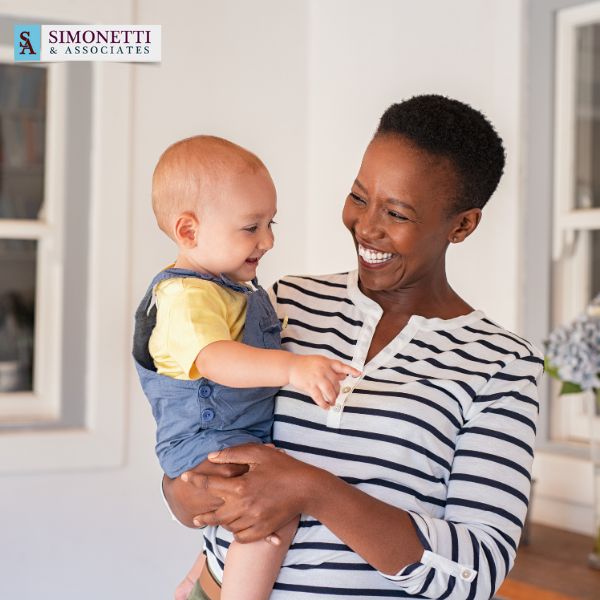 A smiling woman in a striped shirt holds a happy baby in a well-lit room. The Simonetti & Associates logo, known for their trusted Nassau Family Lawyer services, is visible in the top left corner.