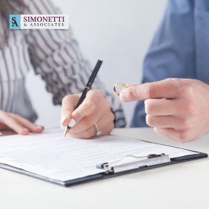 Two people at a desk; one holds a pen over a document on a clipboard, while the other holds a wedding ring. The logo “Simonetti & Associates”—Divorce Lawyer in Southampton—is visible in the top left corner.