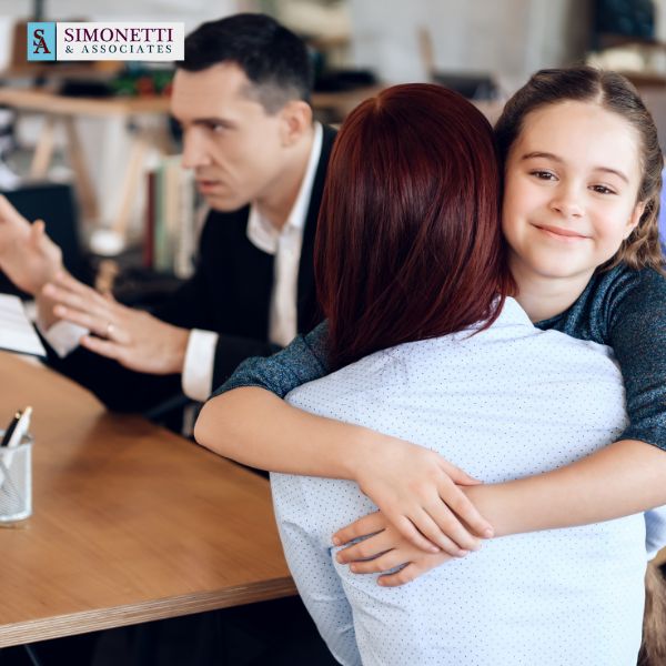 A woman hugs a smiling young girl while a man gestures during a discussion at an office desk. A Simonetti & Associates sign, known for its Suffolk Family Lawyer services, is visible in the background.