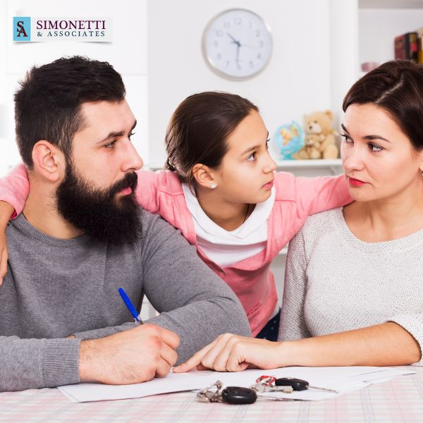A man, woman, and young girl sit at a table with documents and keys, appearing to have a serious conversation. The Simonetti & Associates logo in the top left hints they may be seeking guidance from a Suffolk County family lawyer.