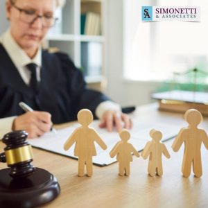 A Nassau family lawyer writes at a desk with wooden family figures, a gavel, and legal papers; Simonetti & Associates logo is visible in the background.