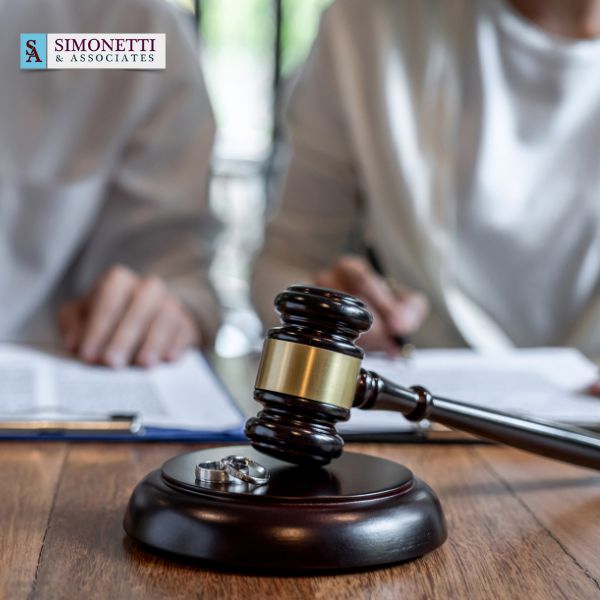 A gavel and wedding rings rest on a table with two people reviewing documents in the background; the Simonetti & Associates logo is visible, highlighting your trusted Huntington NY divorce lawyer.