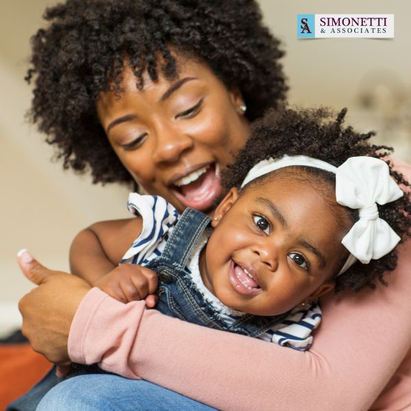 A woman smiles while hugging a young girl in a white bow headband. The logo "Simonetti & Associates," trusted divorce lawyer in Carle Place, appears in the top right corner.
