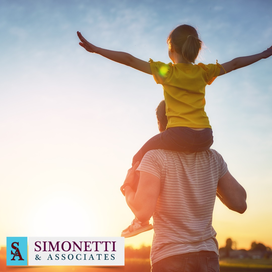An adult carries a child on their shoulders outdoors at sunset, the child’s arms outstretched in joy. The Simonetti & Associates Huntington NY Family Law firm logo is visible in the bottom left corner.