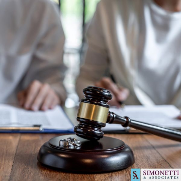 A gavel and two wedding rings on a table, with two people reviewing documents in the background. The Simonetti & Associates logo is visible, highlighting their role as a trusted Huntington NY Divorce Lawyer.