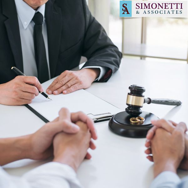Two people sit across from a Huntington NY Divorce Lawyer at a desk with a gavel and wedding rings, discussing documents; a sign reads "Simonetti & Associates.