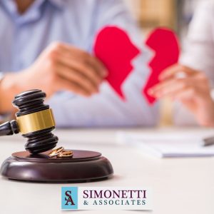 A gavel and two wedding rings rest on a table, with a person holding a broken heart shape in the background; Simonetti & Associates logo visible, highlighting the support of a Suffolk County Family Lawyer.