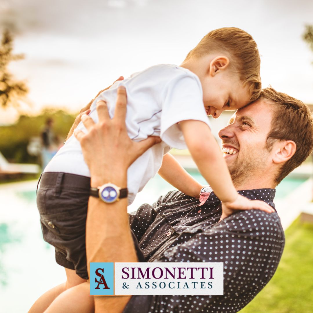 A man smiles while lifting a young boy outdoors near a pool, highlighting family moments. The Simonetti & Associates logo and "Long Island Family Lawyer" are displayed at the bottom of the image.