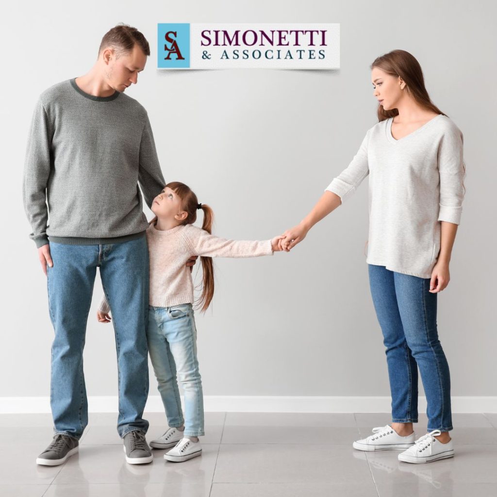 Mother and father stand apart holding hands with their young daughter, who looks up at her father; the