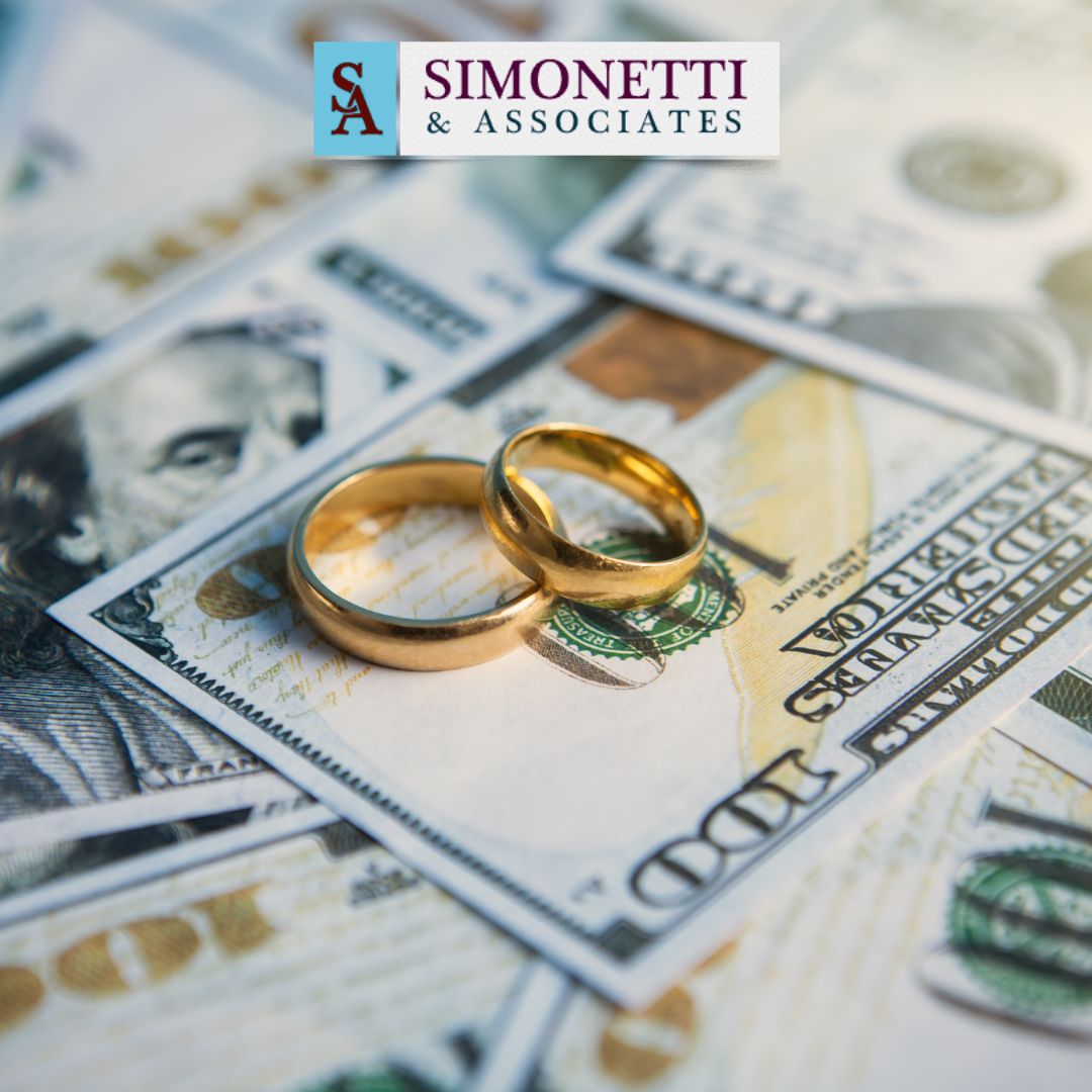 Two gold wedding rings rest on scattered U.S. hundred dollar bills, with a Simonetti & Associates logo in the background, symbolizing the financial aspects handled by a divorce lawyer on Long Island.
