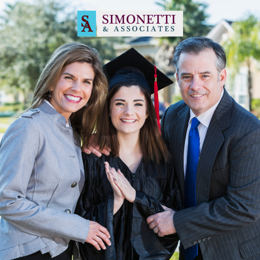 Three people pose outdoors in front of a Simonetti & Associates sign; a young woman wears a graduation cap and gown, flanked by two adults in business attire.