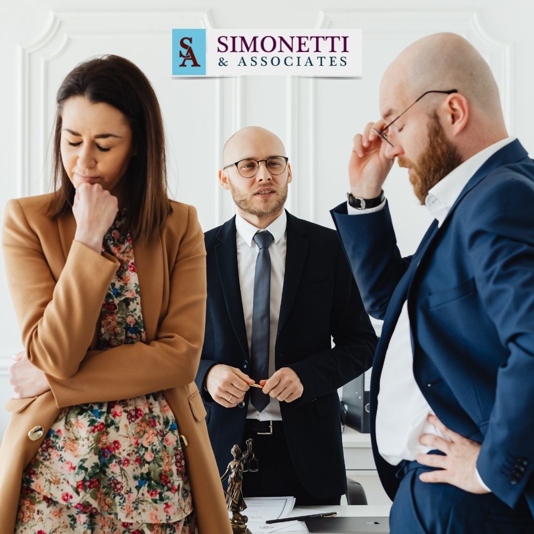 Three people in business attire stand in an office; one woman appears upset, one man looks frustrated, and another man stands between them. The sign reads "Simonetti & Associates.