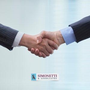Two people in business suits shake hands in front of a blurred office background, representing a successful partnership, with the Simonetti & Associates logo shown at the bottom—trusted advisors for high asset divorce on Long Island.